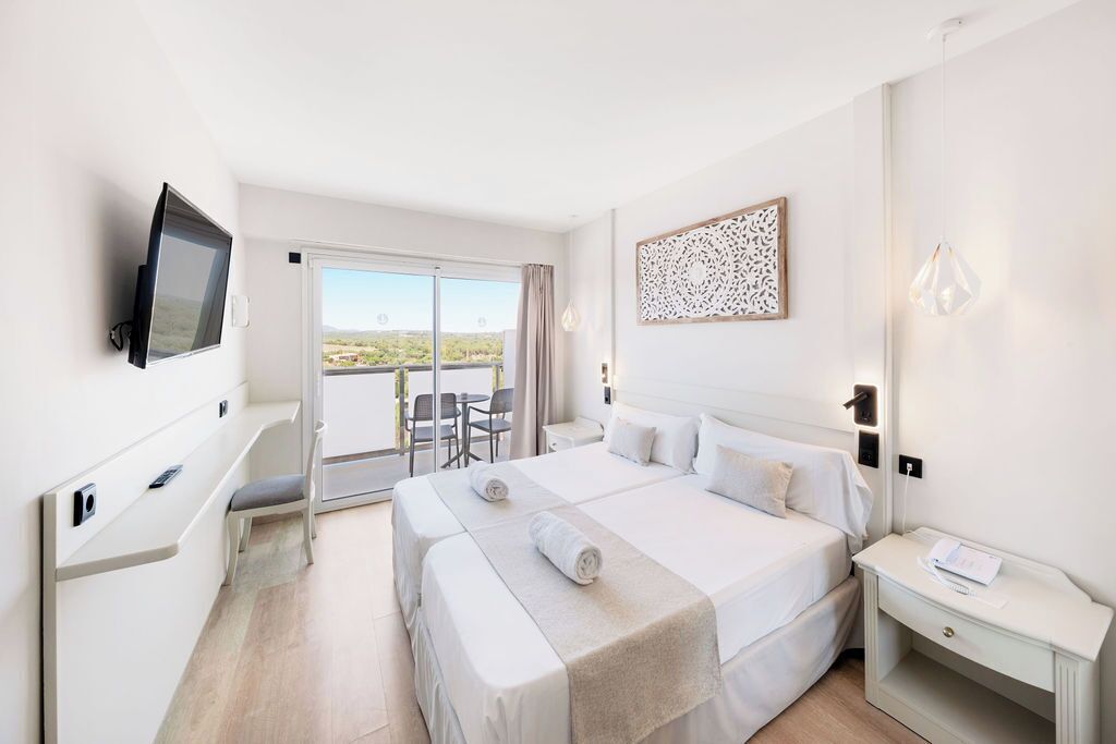 A hotel room with a large bed, a TV mounted on the wall, a balcony with a view, and a small table with a lamp.