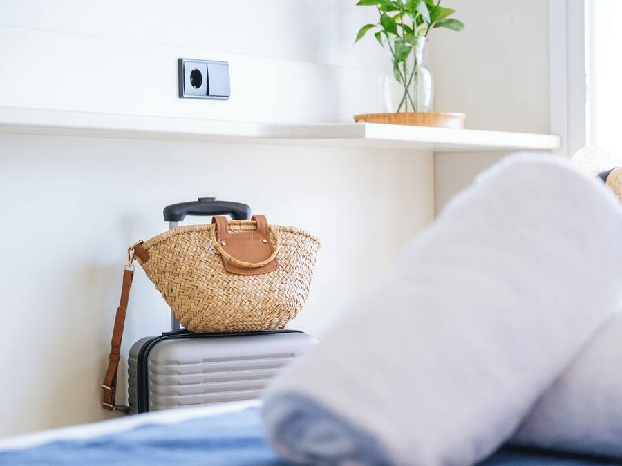 A bed with rolled-up white towels, a suitcase, and a woven handbag on a white nightstand.