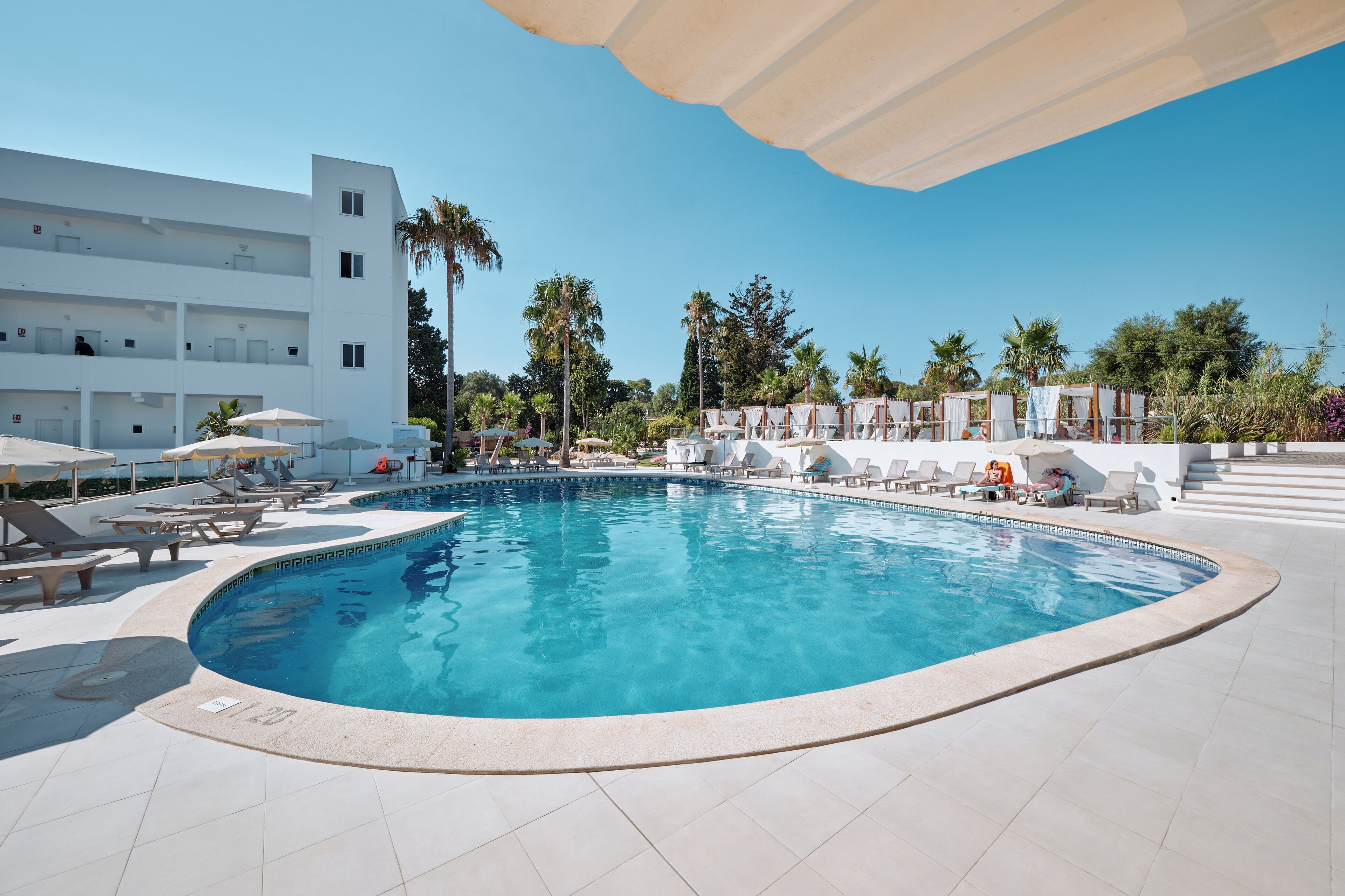 A swimming pool with lounge chairs and umbrellas, surrounded by palm trees and a white building.