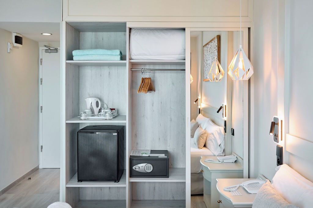 Una habitación de hotel moderna con vestidor, minibar, cama con ropa blanca y luminarias colgantes.