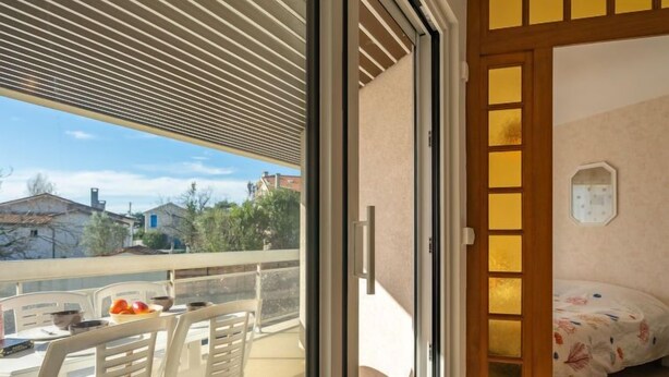 Ein Balkon mit Tisch und Stühlen, ein Bett mit blumigem Überwurf und Blick auf Häuser und Bäume.
