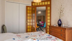 Ein Schlafzimmer mit einem Bett, einer Holzkommode, einer Vase mit Blumen und einem Essbereich, der durch eine offene Tür sichtbar ist.