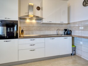 Une cuisine moderne avec des armoires blanches, une hotte de cuisinière en acier inoxydable, un four encastré, une machine à café et une poubelle.