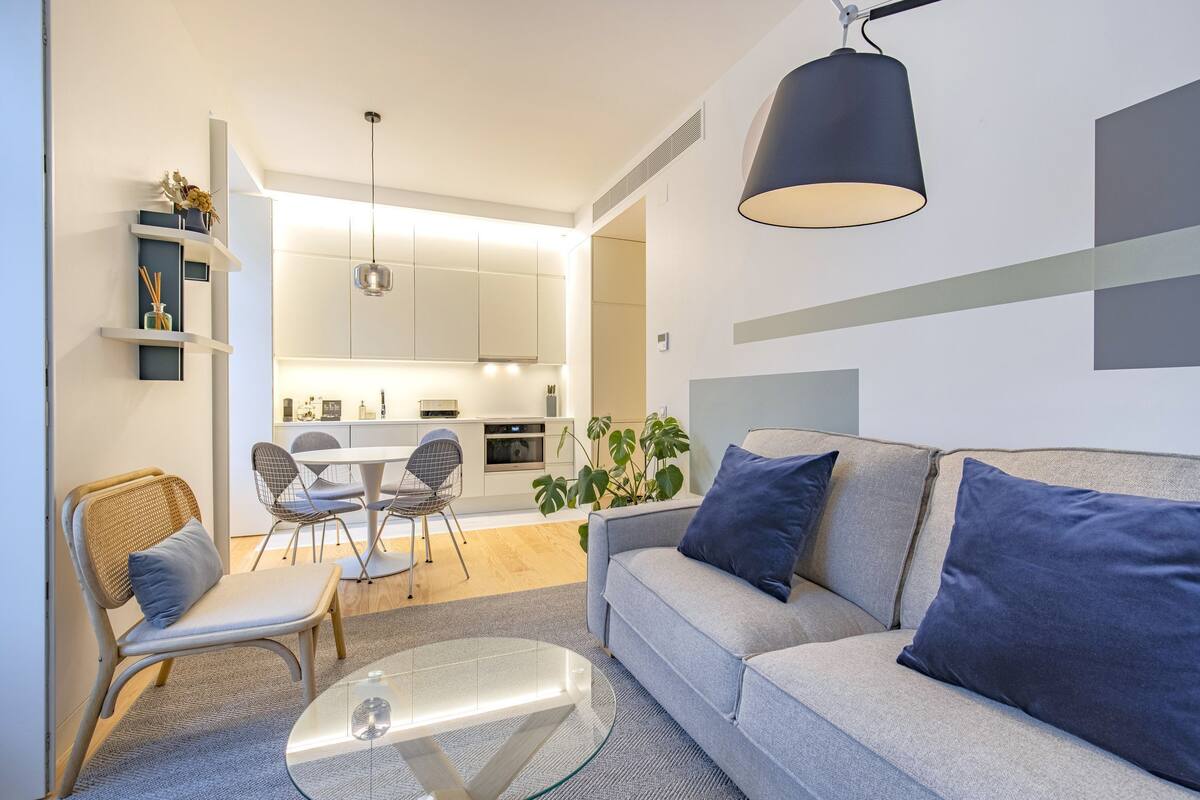 A modern living room with a glass coffee table, a sofa with blue cushions, a wicker chair, and a dining area with a table and chairs.