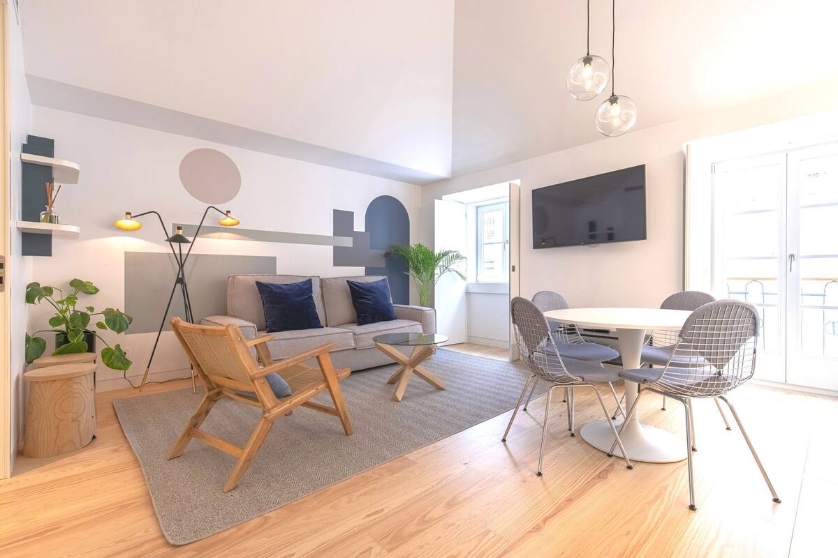 A modern living room with a sofa, coffee table, and chairs. There is a TV mounted on the wall, a plant, and a bookshelf.