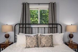 A bed with a headboard, two bedside tables with lamps, and a window with curtains. - Mid-Way on 3rd, Sleeps 10, Sauna and Firepit with tow living rooms and fenced back yard (Kalispell)