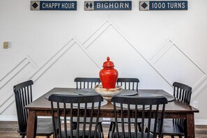 A dining table with four chairs, a red vase centerpiece, and a wall with three signs reading 'CHAPPY HOUR,' 'BIGHORN,' and '1000 TURNS.' - Mid-Way on 3rd, Sleeps 10, Sauna and Firepit with tow living rooms and fenced back yard (Kalispell)
