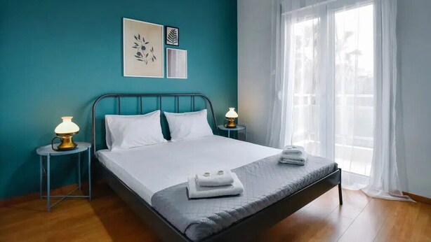 A neatly made bed with white linens and folded towels, a teal-colored wall with framed artwork, and a window with sheer curtains.