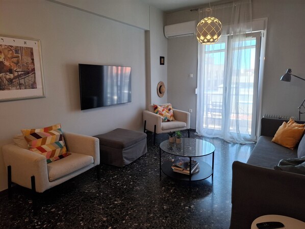 A modern living room with a flat-screen TV, two sofas, a coffee table, and a large window with sheer curtains.