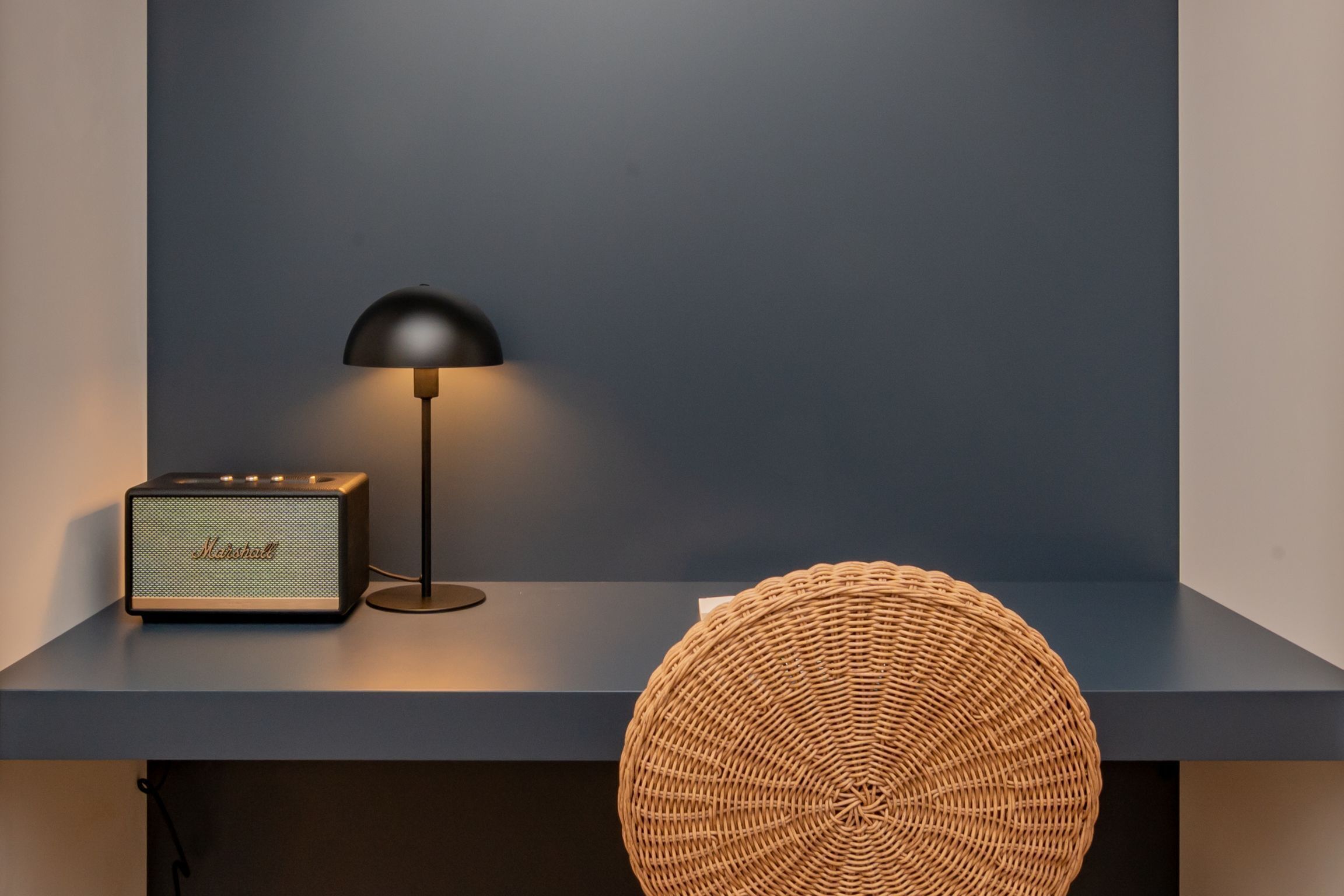 A modern room with a dark blue wall, a black lamp, a vintage-style radio, and a woven round pouf.