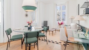 A modern living room with a glass dining table, green chairs, a white sofa, and a large window.
