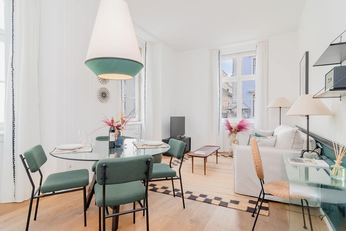 Una sala de estar moderna con una mesa de comedor de vidrio, sillas verdes, un sofá blanco y un ventanal grande.