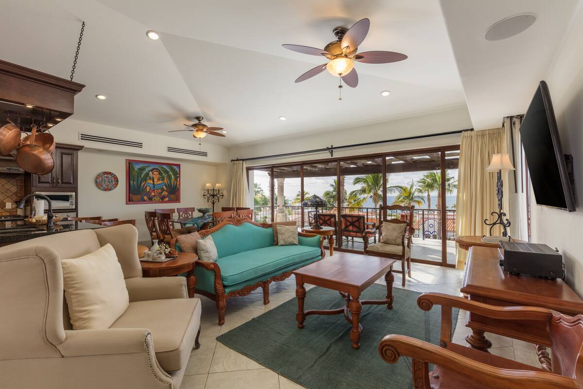 Een ruime woonkamer met een bank, salontafel en stoelen, een keuken met koperen pannen, en een balkon met uitzicht op palmbomen.