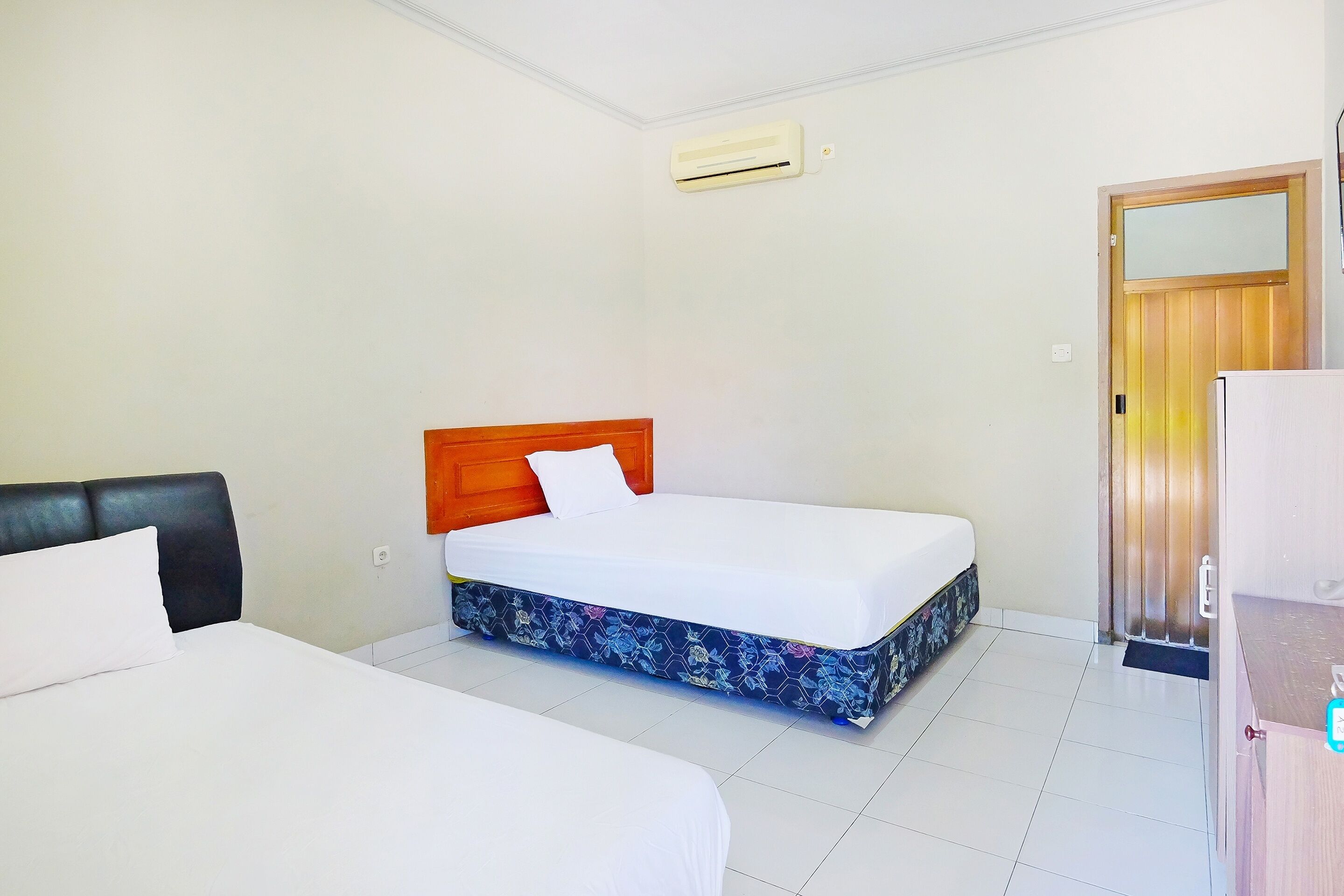 A hotel room with two beds, a wooden headboard, a white mattress, a floral-patterned mattress cover, a white pillow, a wooden door, and a white air conditioning unit.