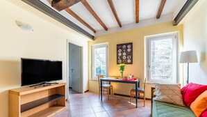 A living room with a TV on a wooden stand, a dining table with chairs, a sofa with cushions, and a window with a view. - GuestHost - Two-room apartment located on the second floor of a building, without lift, located in the heart of the historic center of Parma. The property enjoys a central location (ZTL), in a picturesque, popular and lively district, near the Don Gnocch (Parma)