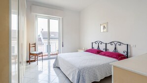 Un dormitorio con cama, una silla, una mesita de noche, un cuadro en la pared y una puerta corrediza de vidrio que da a un balcón exterior.