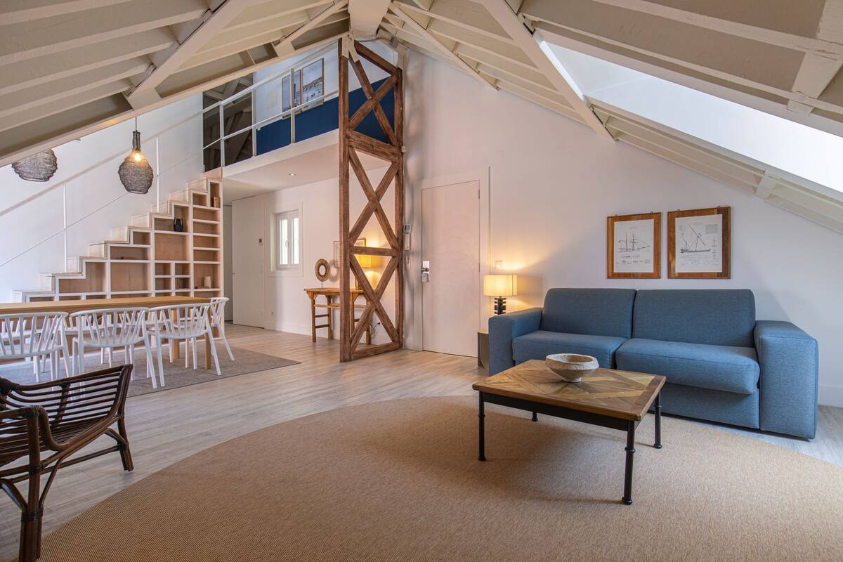 Un loft espacioso con techo alto, vigas de madera expuestas, un sofá azul, una zona de comedor con sillas blancas y una escalera que lleva a un nivel superior.
