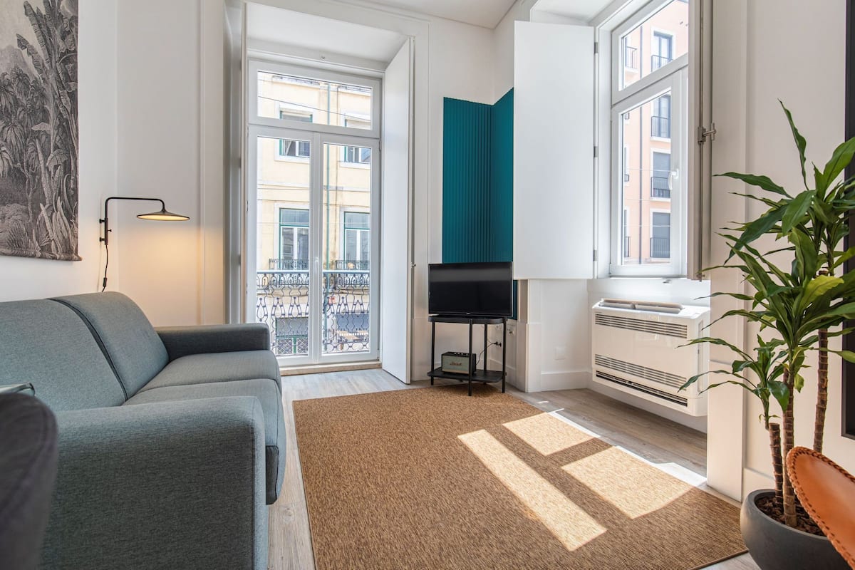 Una sala de estar moderna con un sofá gris, un televisor de pantalla plana sobre un mueble bajo, un ventanal con vistas a edificios y una planta en maceta.