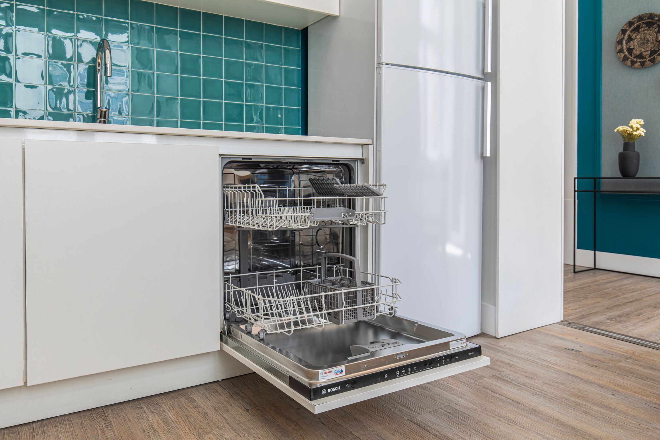 Une cuisine moderne dotée d’un lave-vaisselle Bosch, de meubles blancs, d’un pare-feu turquoise et d’un plancher en bois.