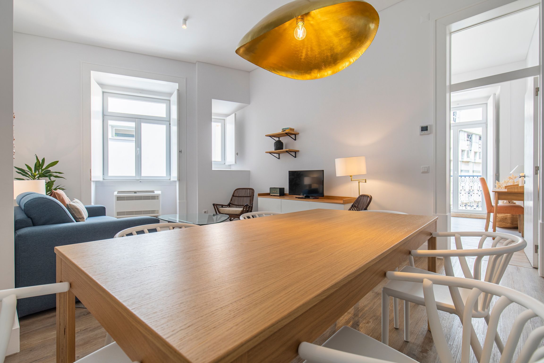 A modern living room with a wooden dining table, white chairs, a blue sofa, and a large window.