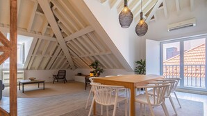 Een ruime kamer met een hoge plafond, een houten eettafel met witte stoelen, een groot raam met uitzicht op een gebouw met rode dakpannen, en een hanglamp.