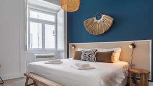 Een slaapkamer met een groot bed, een houten bankje, een nachtkastje en een wandlamp.