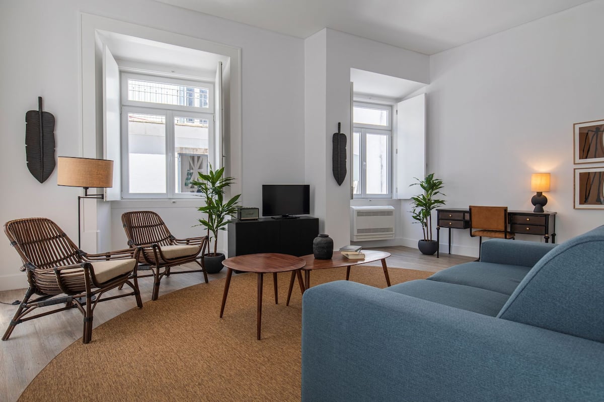 Una sala de estar moderna con un sofá, dos sillones, una mesa de centro y un televisor. Hay plantas en macetas y una lámpara sobre una mesita auxiliar.