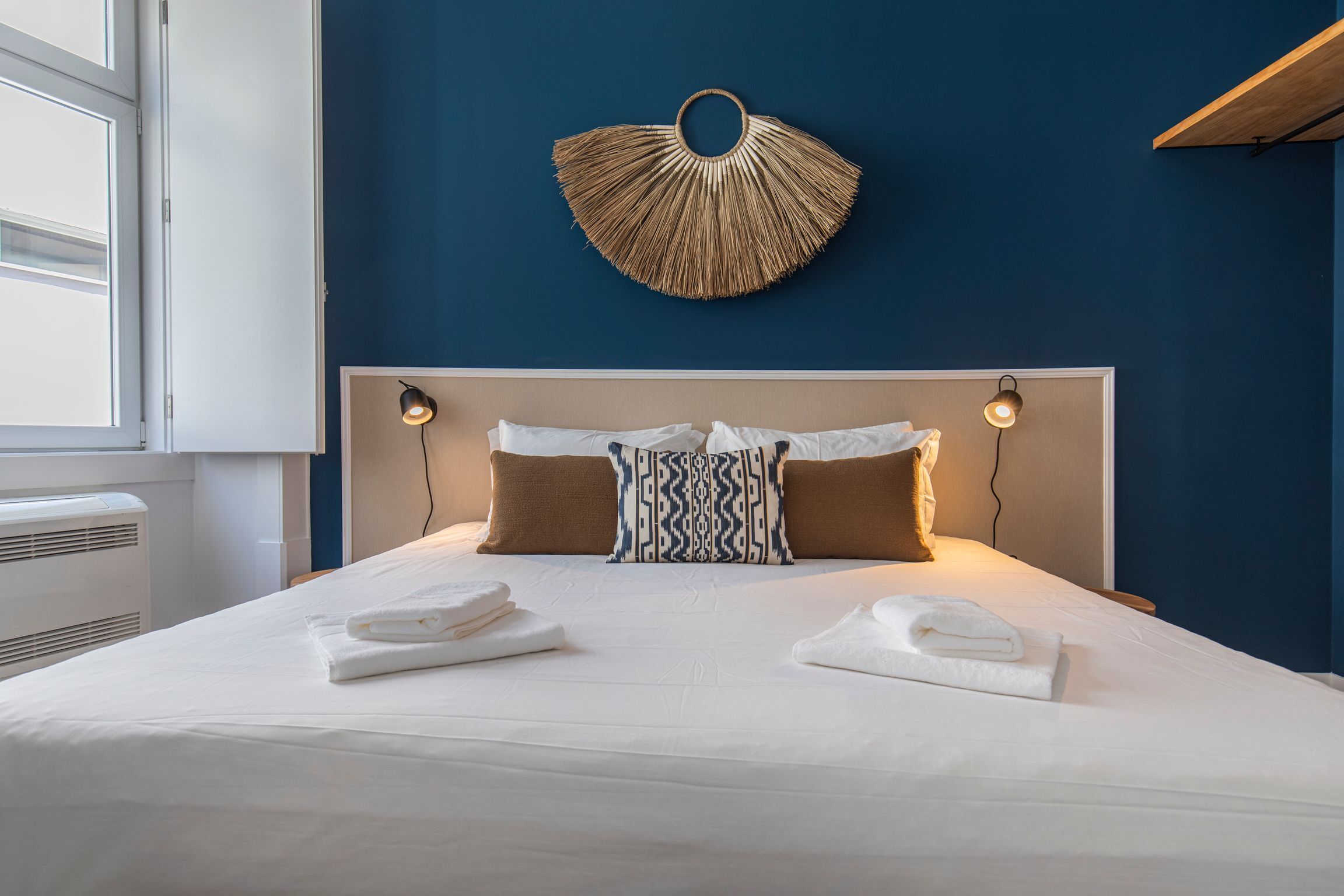 Una cama bien hecha con sábanas blancas, dos toallas dobladas, un cojín estampado y un ventilador de pared.