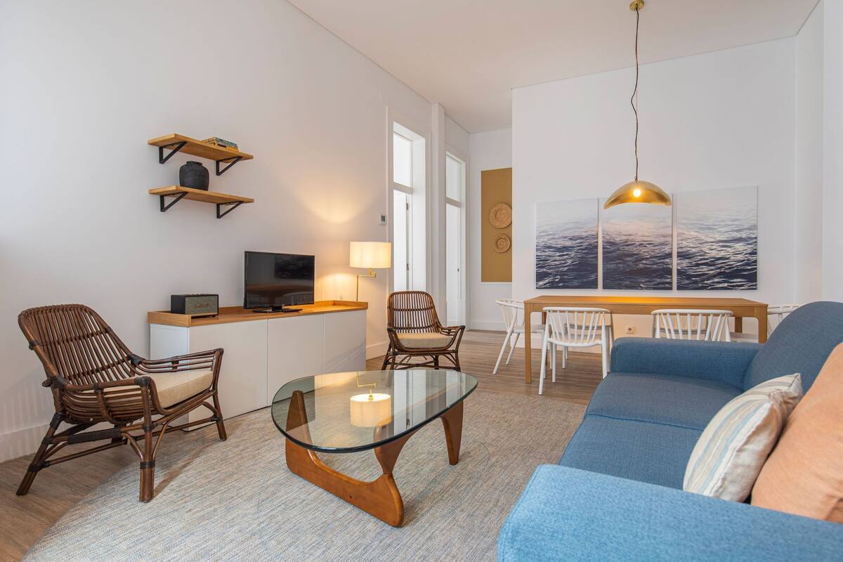 Un salón moderno con una mesa de centro de vidrio, un sofá azul, sillas de mimbre, una zona de comedor con una mesa blanca y un televisor sobre un mueble de madera.