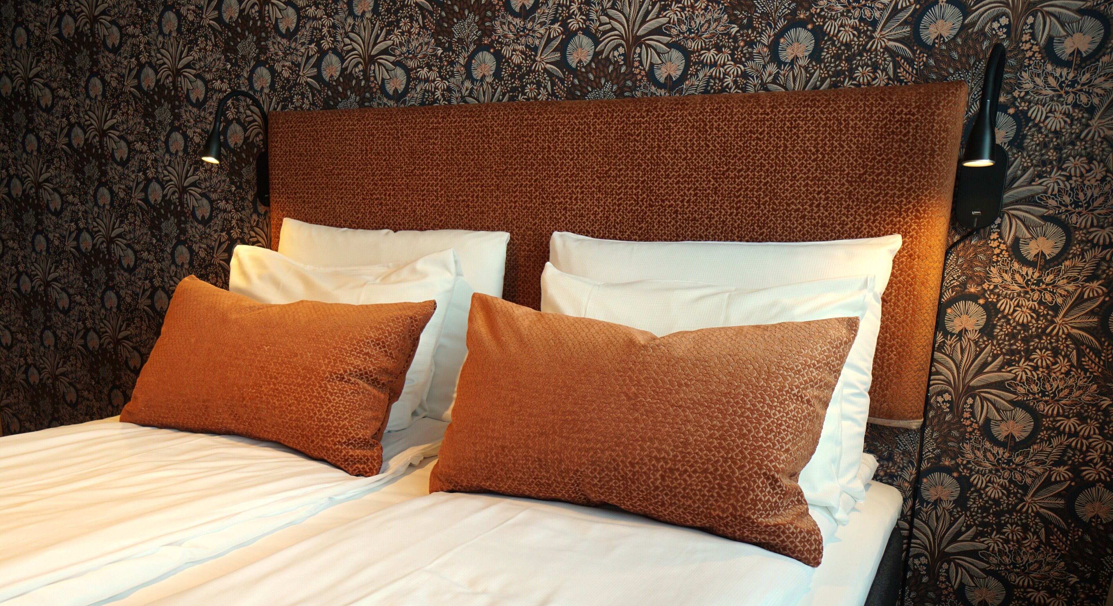 A bed with a patterned headboard, two pillows, and a wall with floral wallpaper.