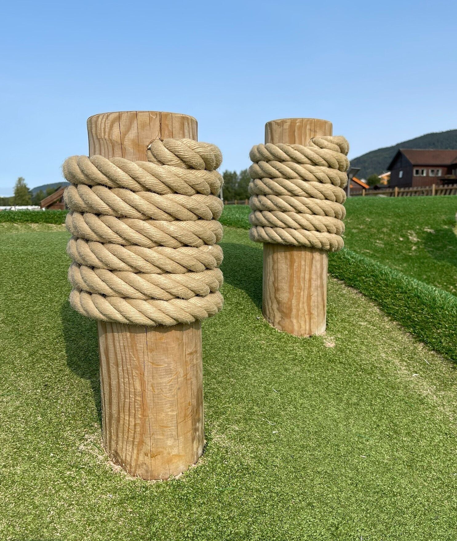 Two wooden posts wrapped with thick rope, set on a grassy field with a house in the background.