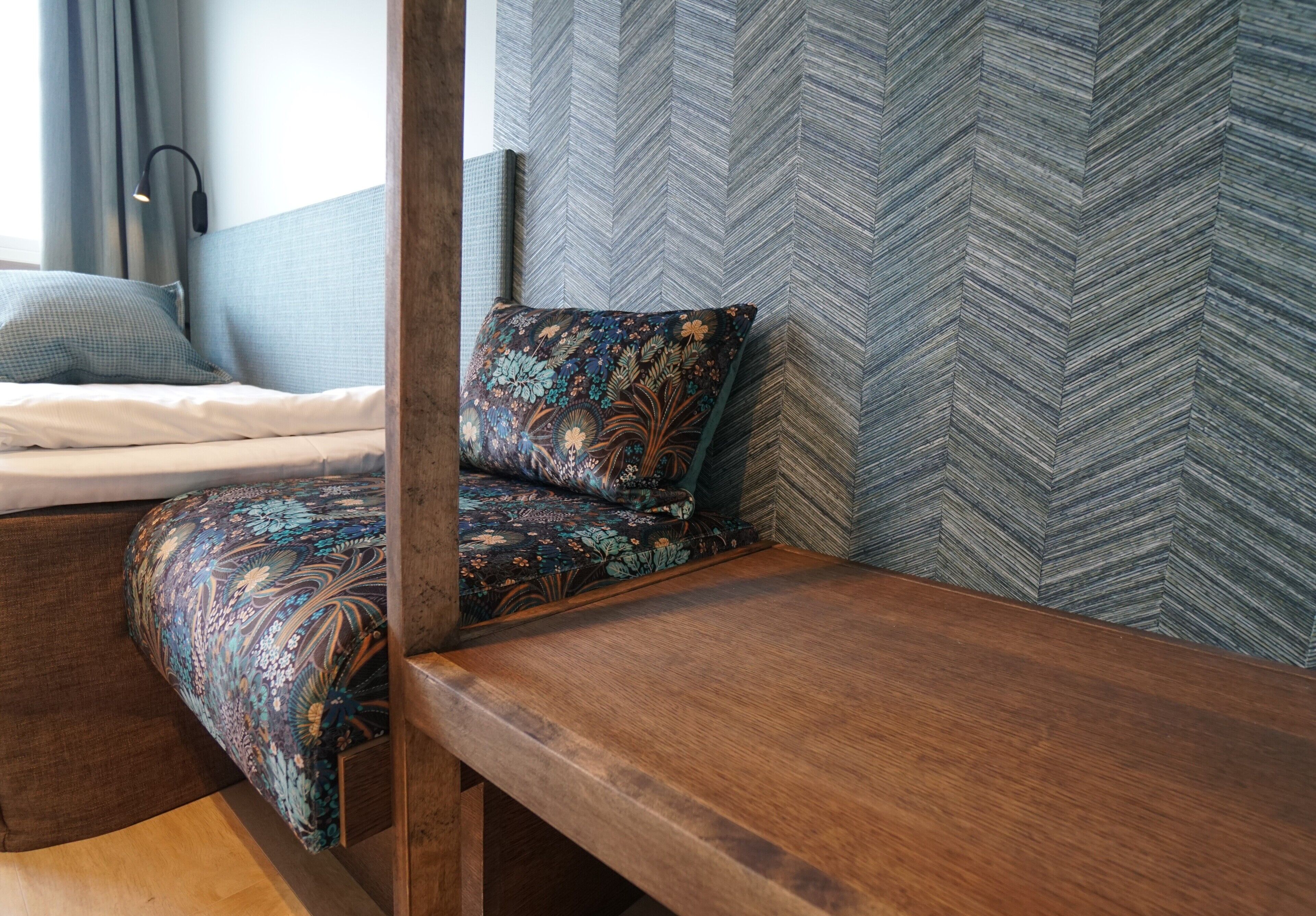 A corner of a room with a bed, a wooden bench, and a patterned wall.