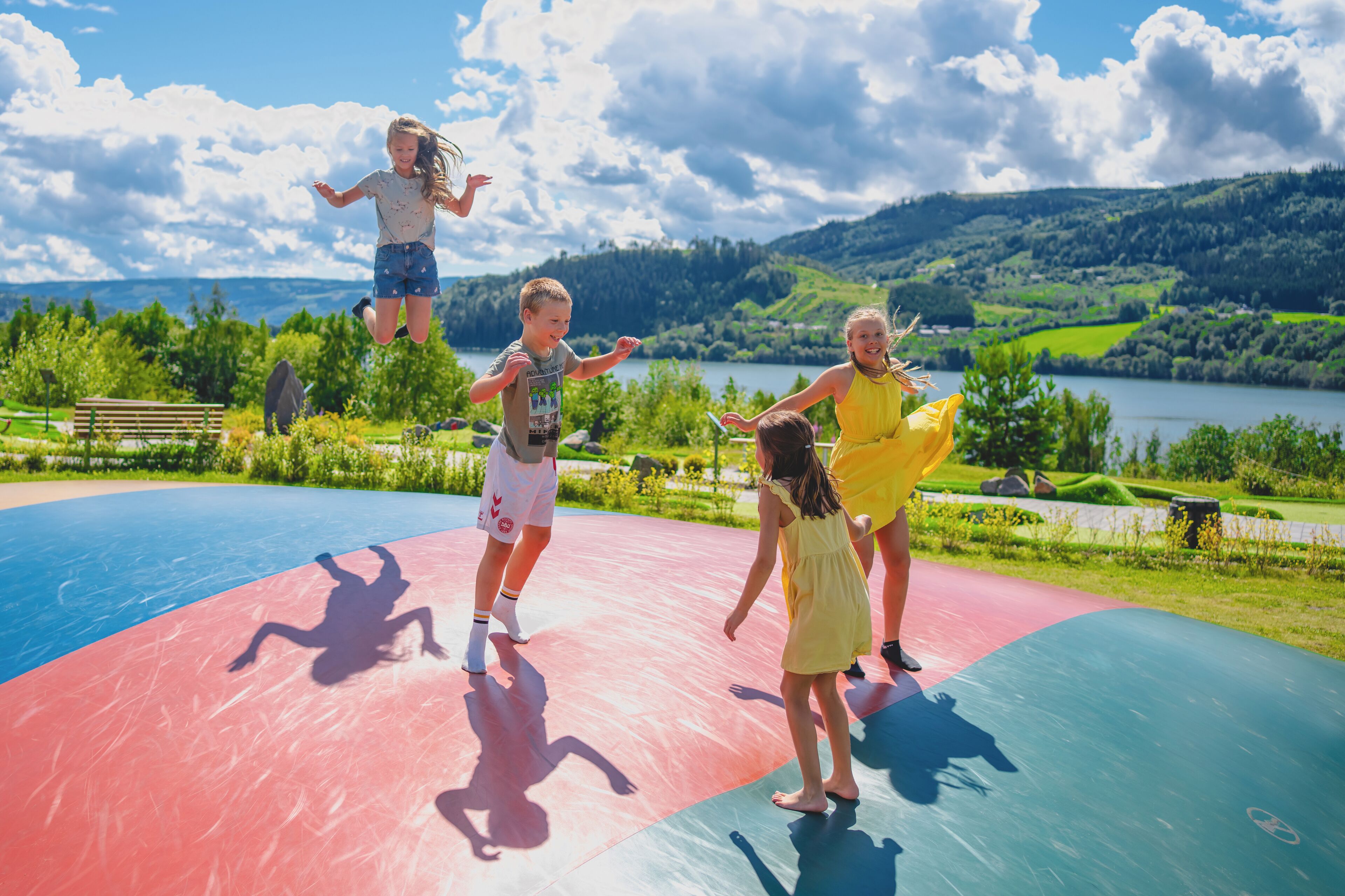 A group of children playing on a colorful outdoor play area with a scenic lake and hills in the background.