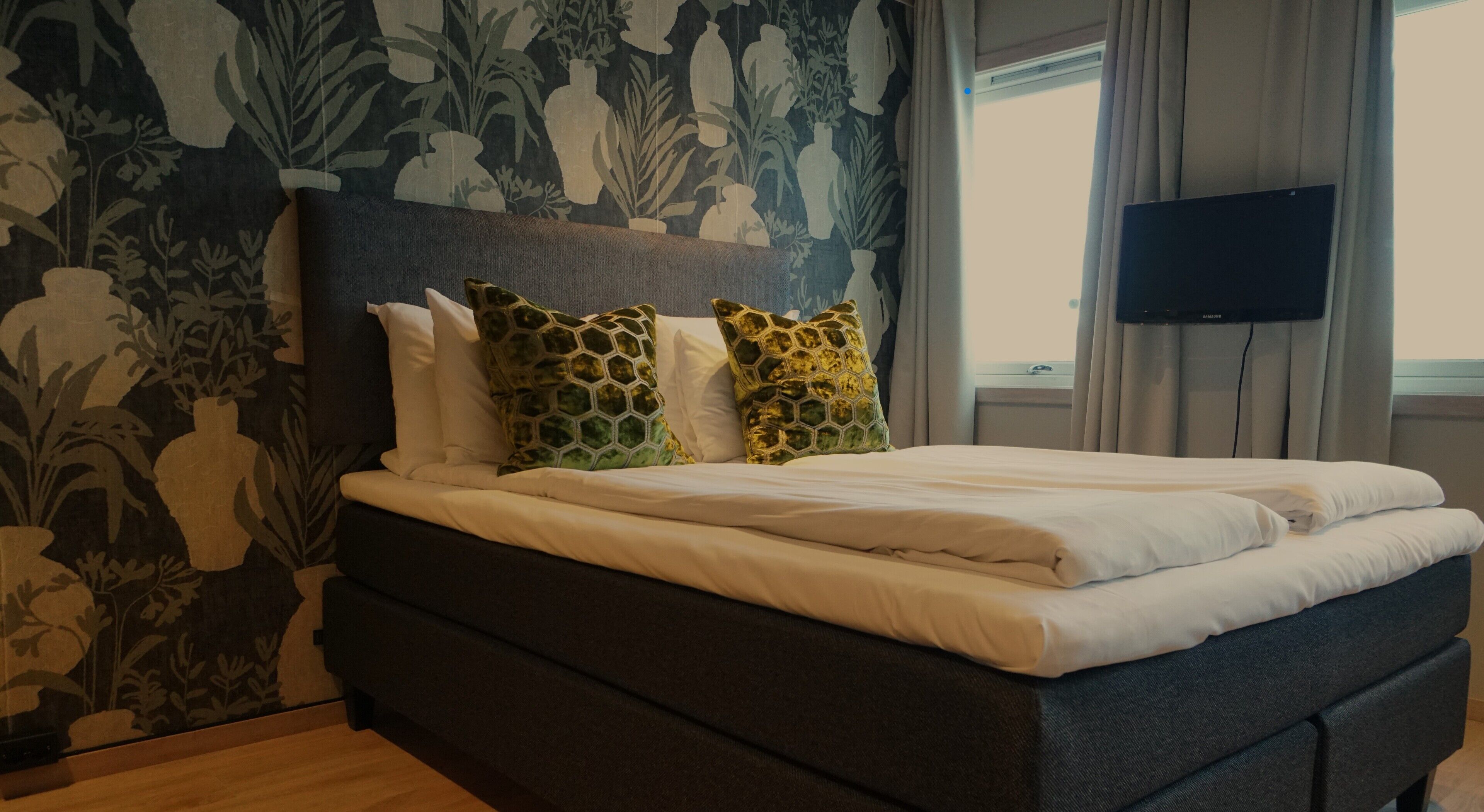 Una cama bien hecha con un cabecero oscuro y cojines decorativos, un televisor montado en la pared y una ventana con cortinas.