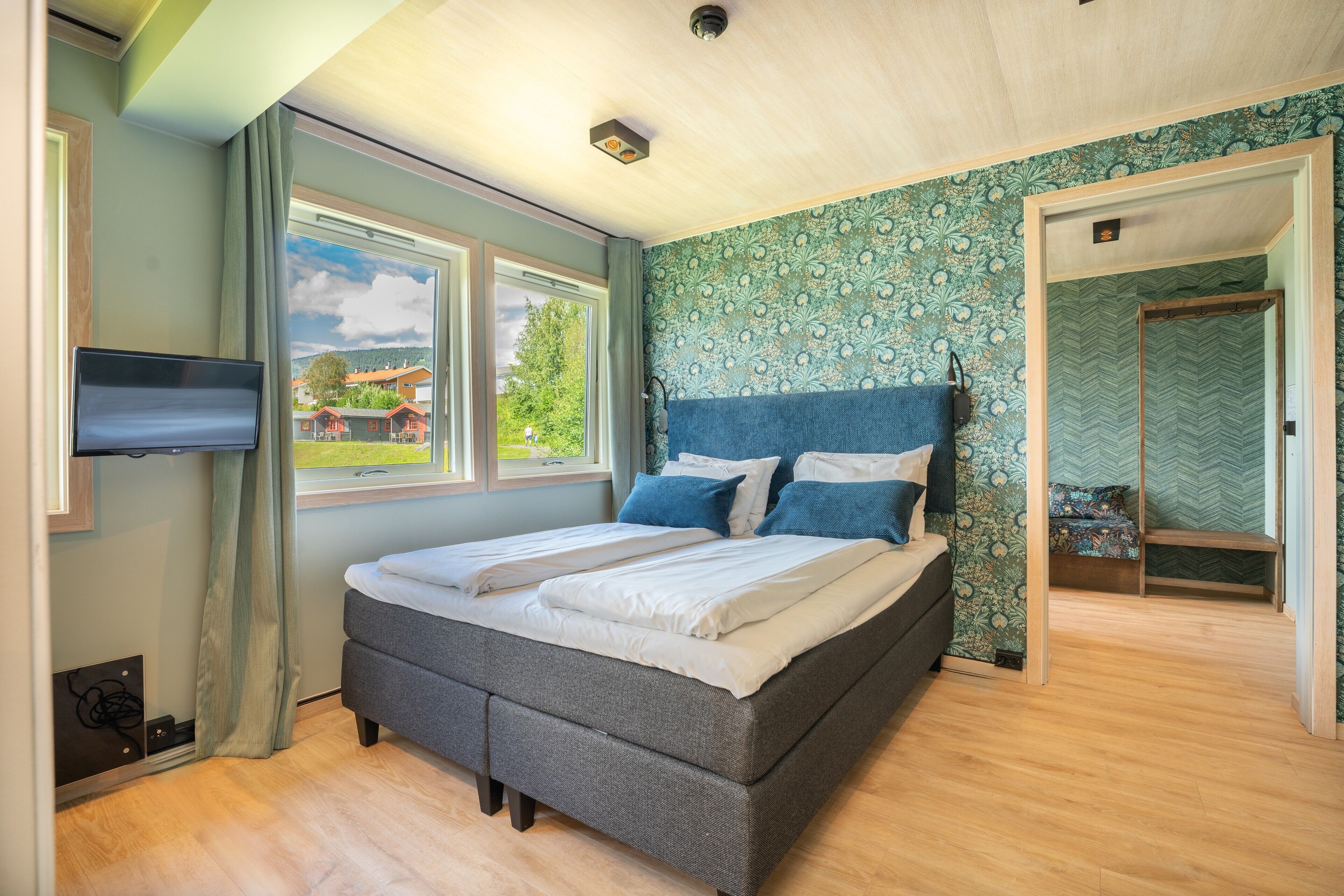Een slaapkamer met een groot bed, een aan de muur bevestigde televisie en een raam met uitzicht op groen en gebouwen.