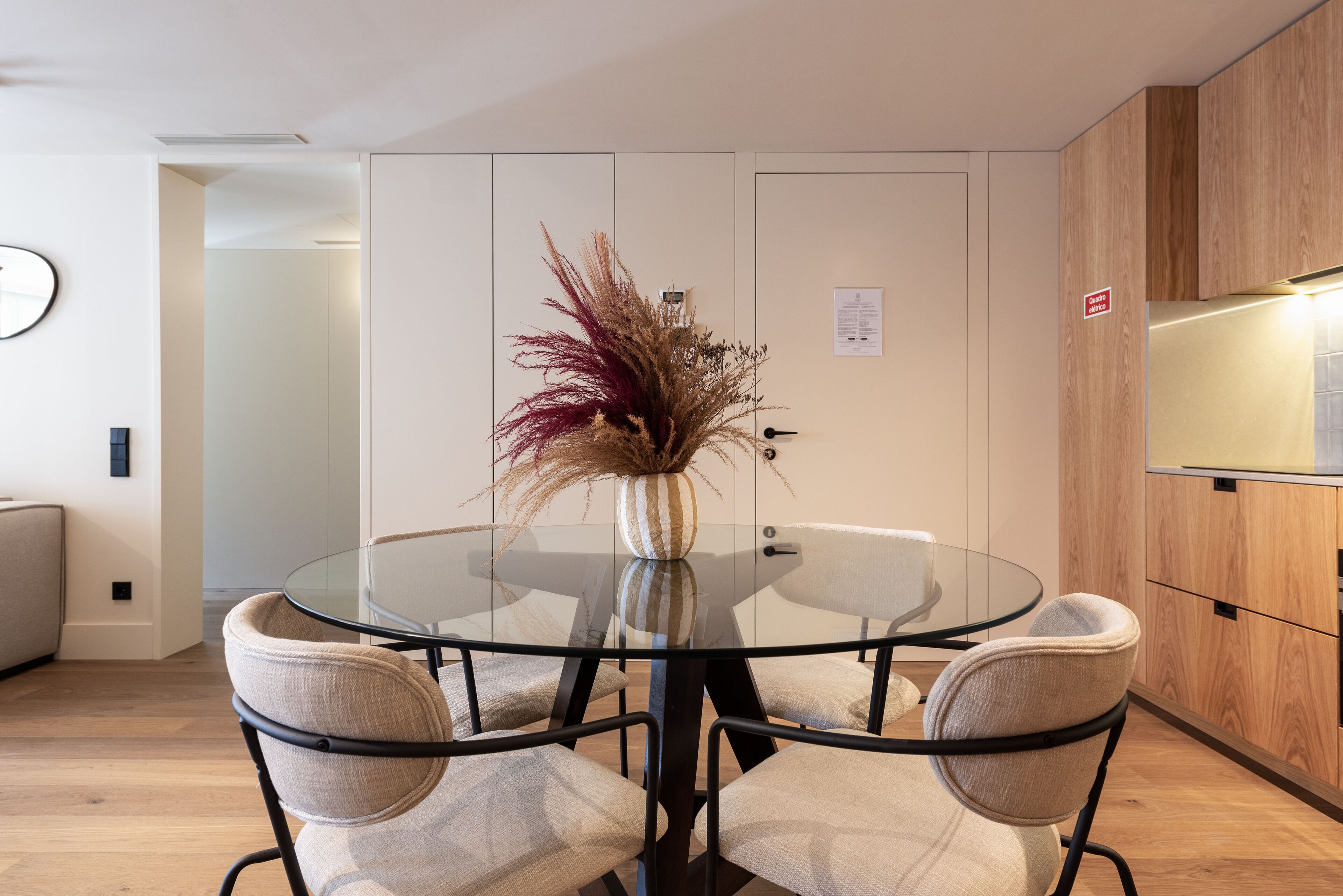 A modern dining area with a glass table surrounded by cushioned chairs, a decorative vase with dried plants, and wooden cabinetry.