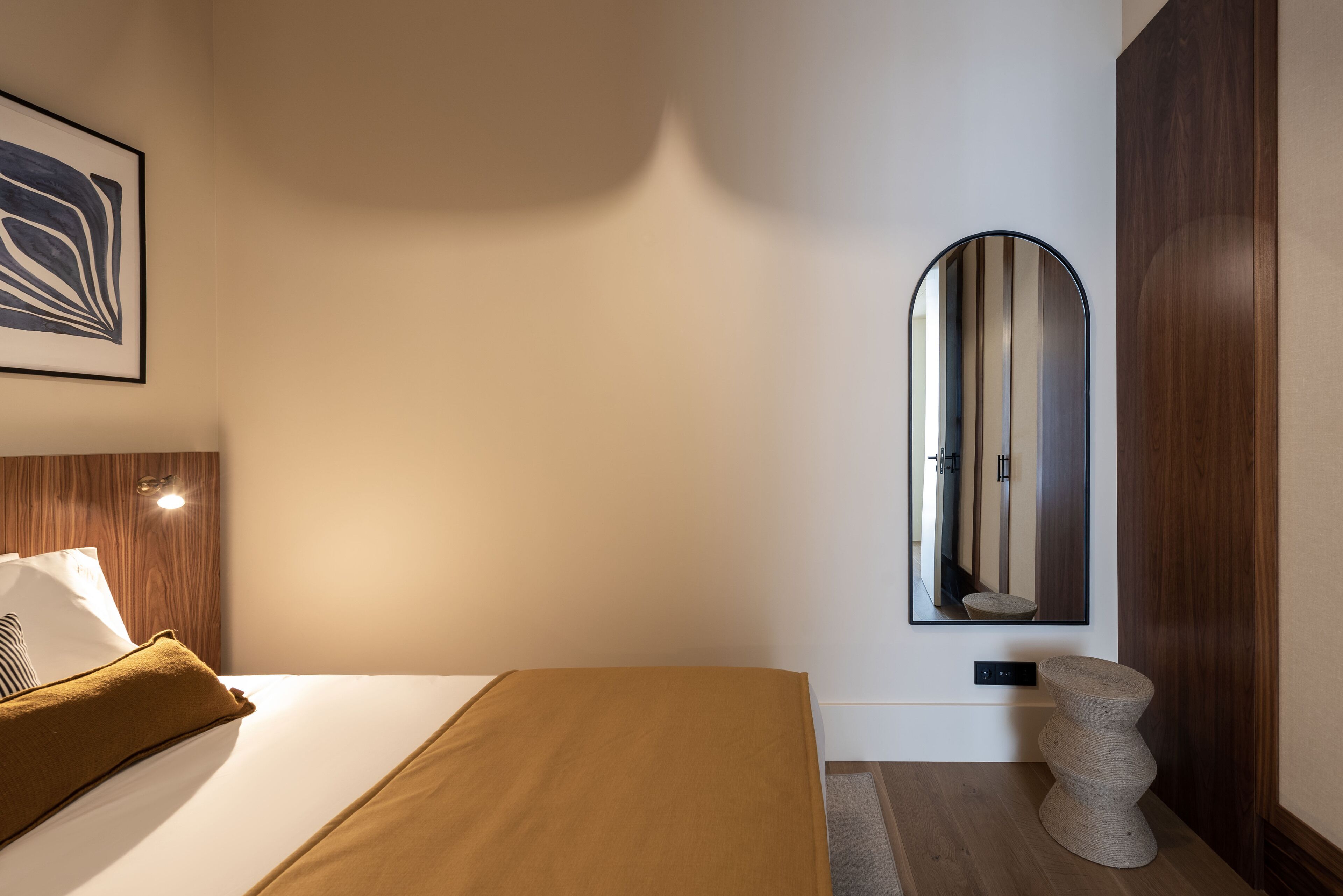 A hotel room with a bed, a wooden headboard, a large mirror, and a stone stool.