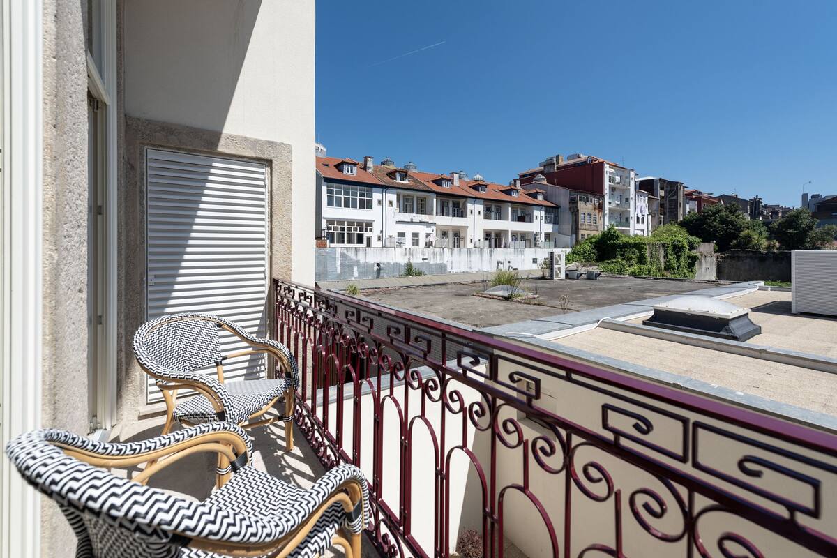 Un balcon avec des fauteuils en osier et une rambarde en métal, donnant sur un quartier résidentiel avec des immeubles et un ciel dégagé.