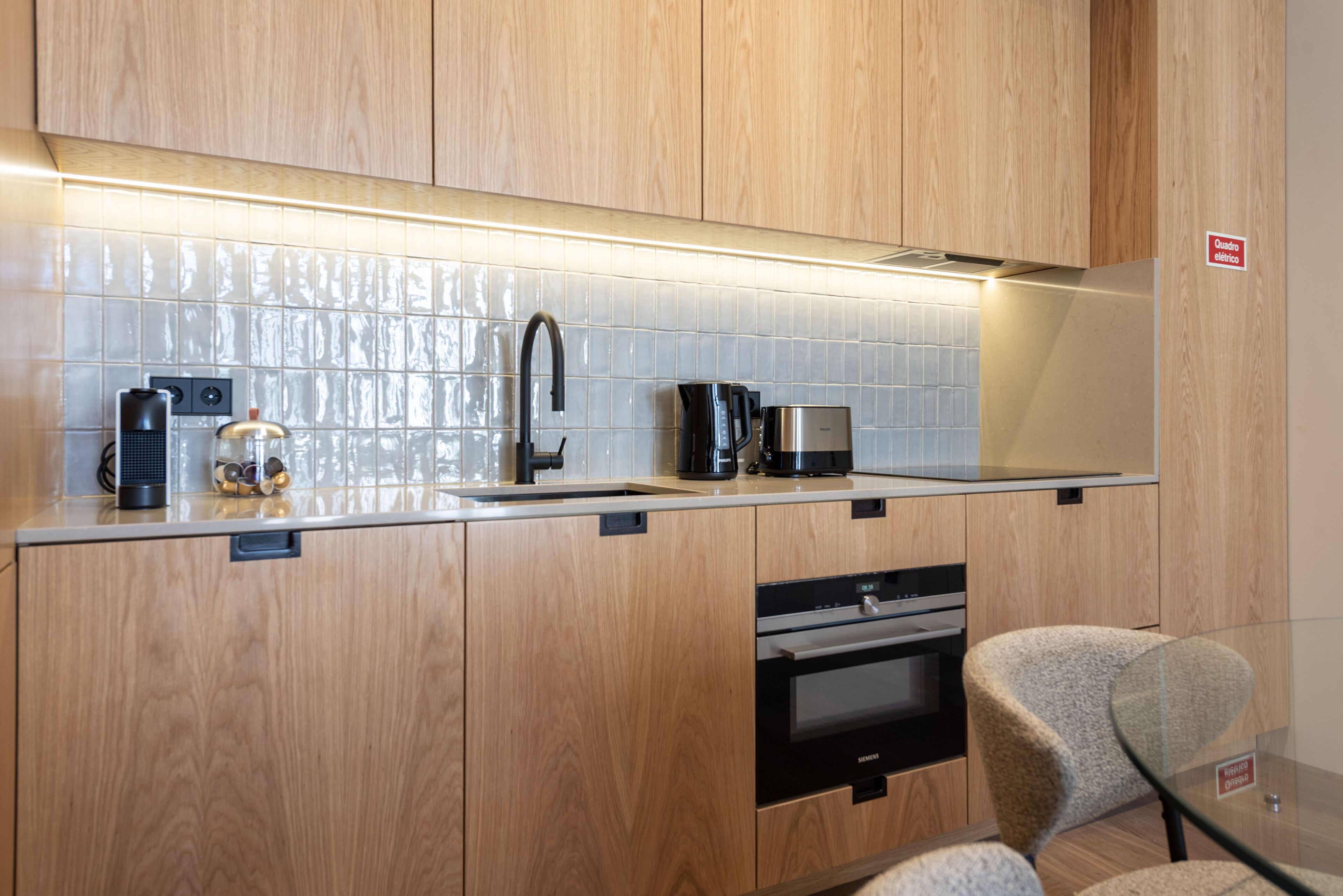 Una cocina moderna con armarios de madera, un lavamanos de acero inoxidable, una cafetera y una mesa de vidrio con una silla.