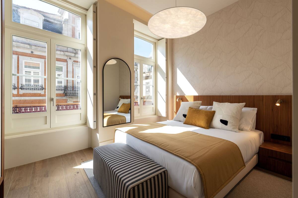 Une chambre d’hôtel moderne avec un grand lit, un banc, un miroir et une vue sur la ville grâce à de grandes fenêtres.