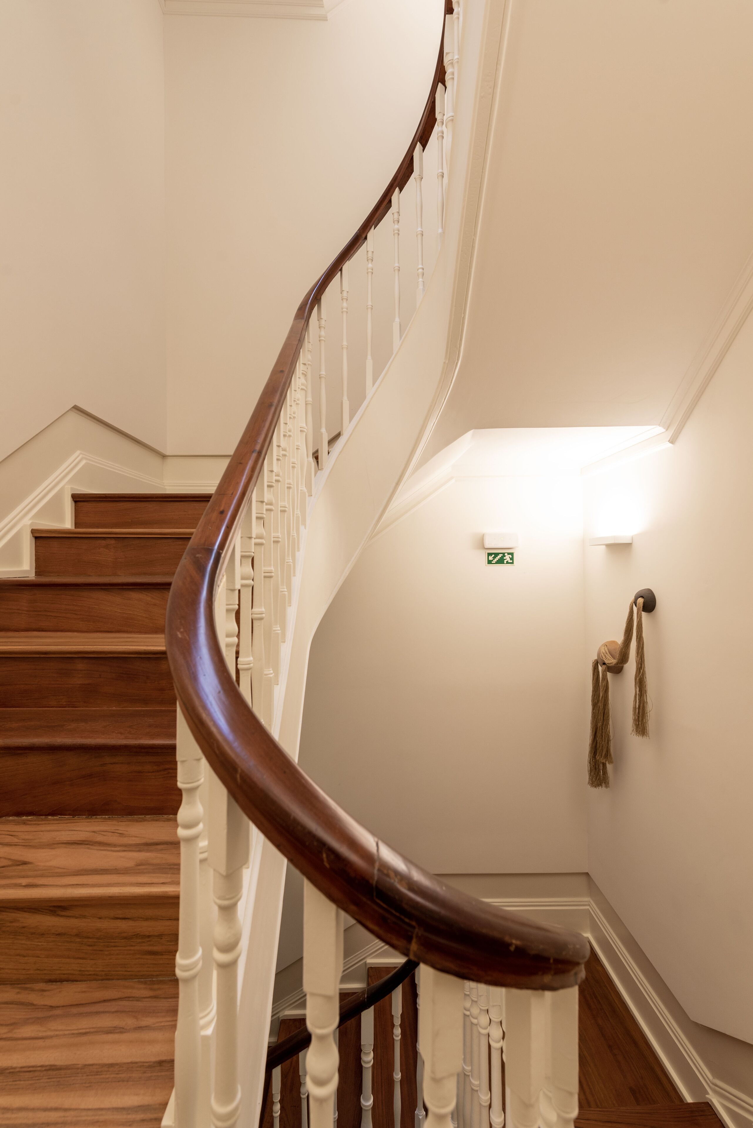 A spiral staircase with wooden steps and white balustrades, leading to a landing with a green exit sign.