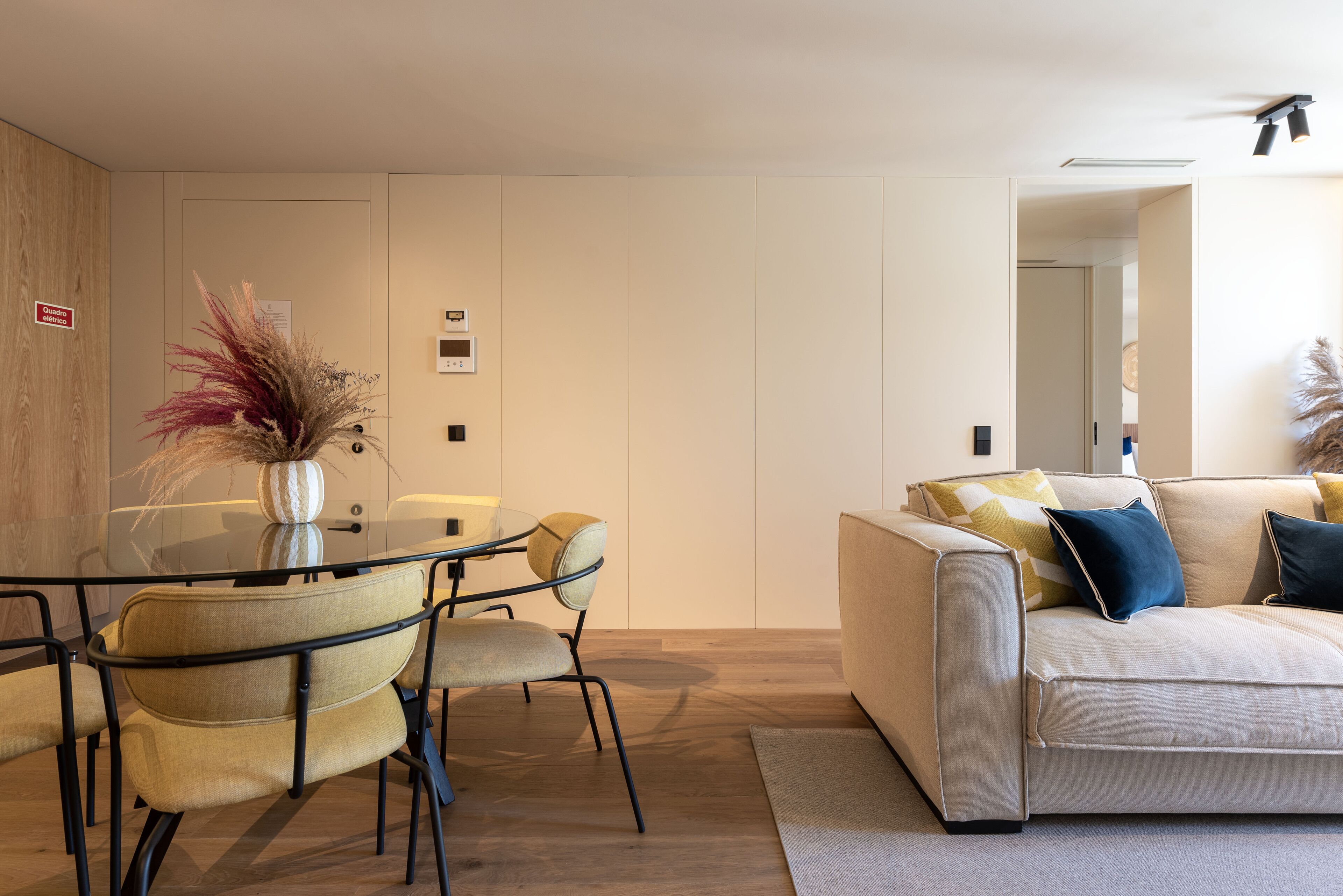 A modern living room with a glass dining table, beige sofa, and matching chairs.