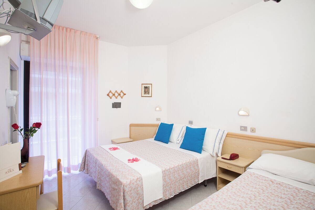 Habitación de hotel con dos camas, un escritorio y una silla. Se observa una ventana con cortinas translúcidas y un florero con flores sobre el escritorio.