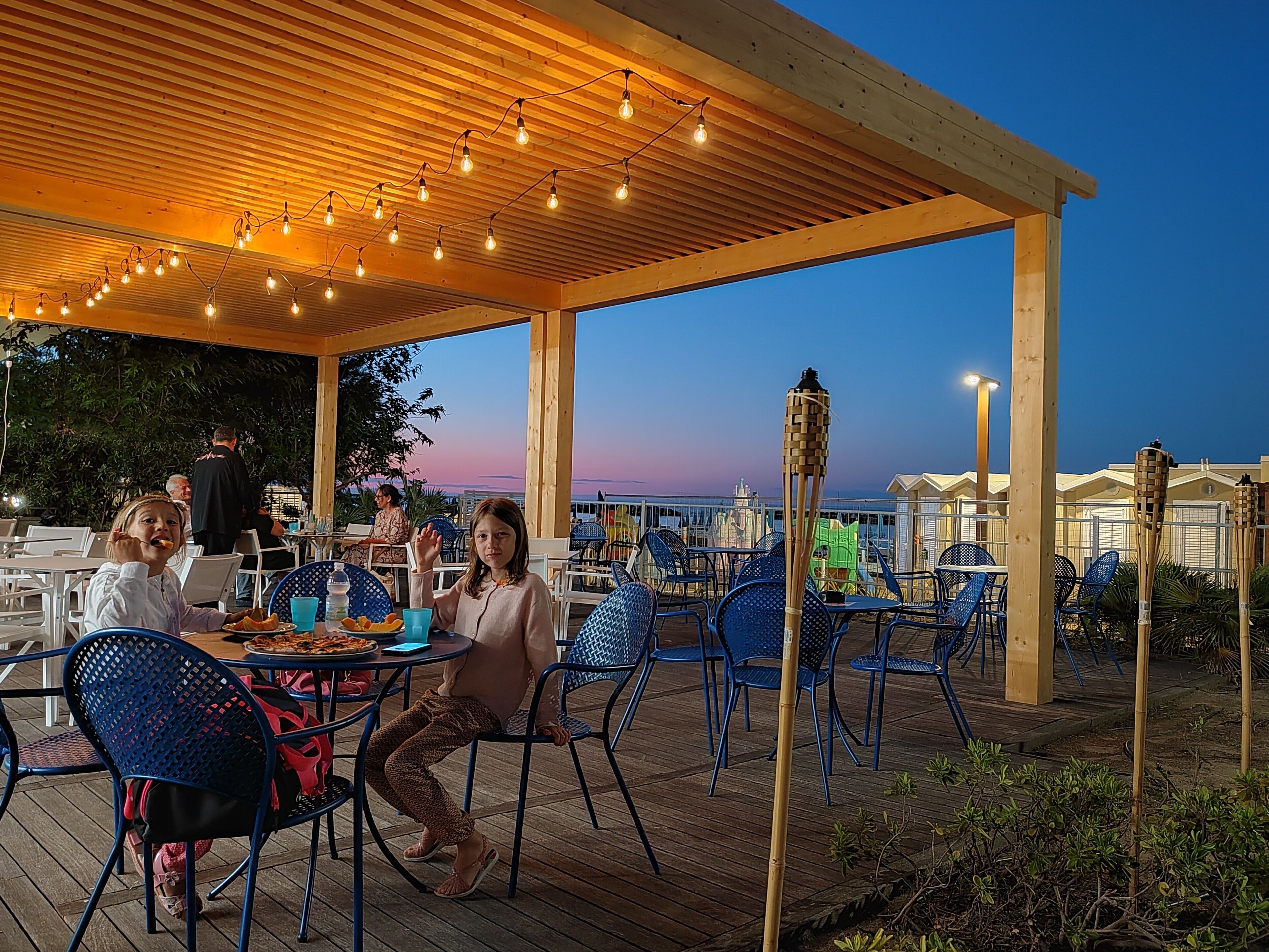 An outdoor patio with wooden tables and chairs, string lights, and a view of the city skyline.