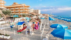 Ein Hotel direkt am Strand mit mehreren Balkonen, einem Spielplatz und zahlreichen Sonnenschirmen.