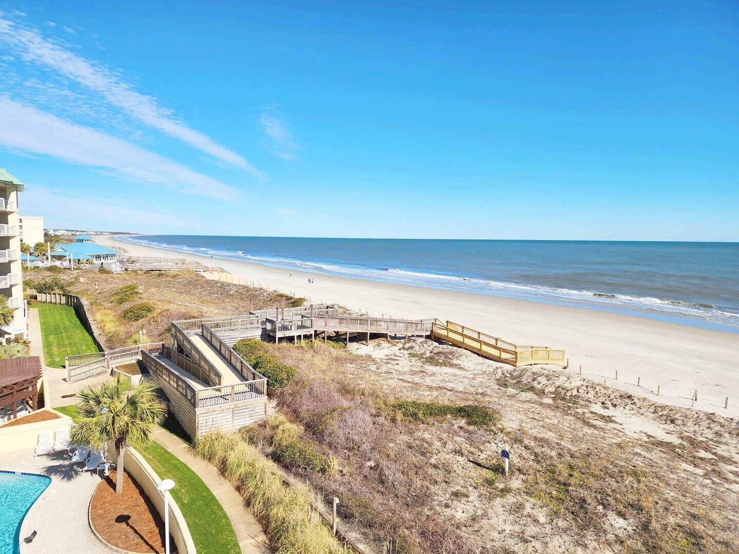 A beach view with a wooden boardwalk, a pool, and a building with a balcony.