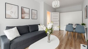 Ett modernt vardagsrum med en grå soffa, ett vitt soffbord och en vas med vita blommor. På väggen hänger inramade tavlor, och det finns en matplats med ett vitt bord och blå stolar. En vit dörr leder in till ett annat rum.