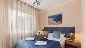 Ein Hotelzimmer mit Bett, Nachttisch, Lampe, Tablett mit Vase und Tassen sowie einem Gemälde einer Küstenlandschaft.
