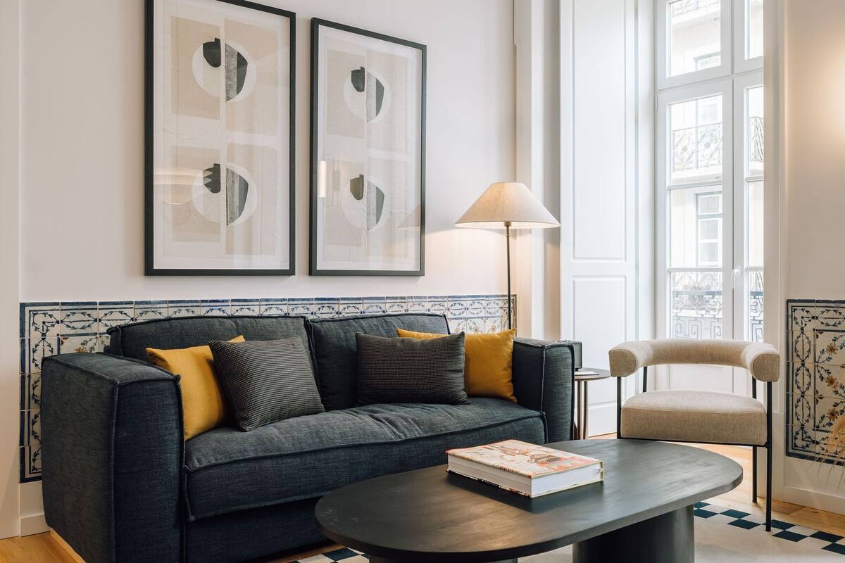 Ein modernes Wohnzimmer mit einem dunkelgrauen Sofa, einem runden Couchtisch mit einem Buch, einer Stehlampe und einer dekorativen Vase mit Trockenpflanzen.