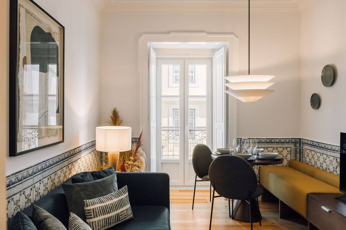 Ein Wohnzimmer mit einer Couch, einem Esstisch mit Stühlen, einer Lampe und Blick auf ein Gebäude durch eine Tür.